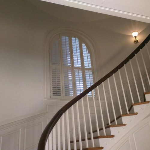White plantation shutters adorning rounded window located in spiral stairwell.